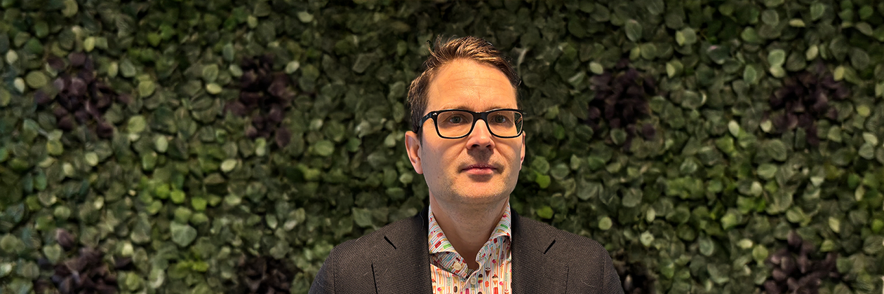 Een persoon in een gestreepte shirt en een donkere blazer staat voor een groene, bladerrijke muur.